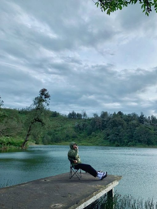 A person sits on a folding chair at the end of a concrete pier extending into a calm lake, surrounded by lush green hills under a cloudy sky.