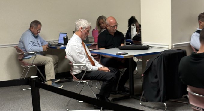 A group of people sit at tables in what appears to be a meeting or conference room, with one man in a white shirt and orange tie in the center foreground looking at his phone, while others work on laptops.