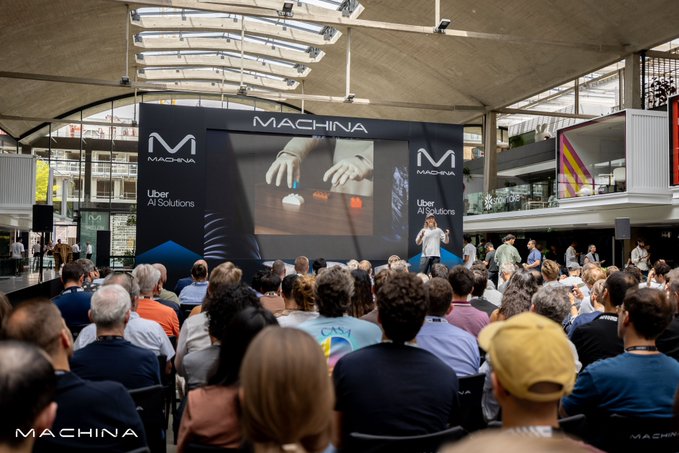 A large audience gathers at an indoor venue for a Machina conference, watching a speaker present on stage with a screen displaying the Machina logo and "Uber AI Solutions" branding alongside an image of robotic hands. The event setting aligns with the post's positive review of Machina's event, suggesting a tech or AI-focused gathering where the presenter likely discussed topics including NEOs.