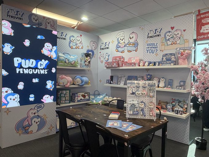 A Pudgy Penguins merchandise display/store setup showing branded banners, plush toys, accessories, and "MADDLE HUGGIES" merchandise on shelves.