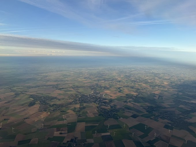Aerial photograph of a vast green agricultural landscape with scattered towns, wind turbines, and power lines, taken from an airplane window with a blue sky overhead.