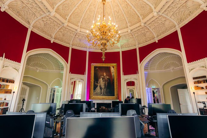 An ornate circular room with red walls, white decorative ceiling, a large chandelier, a classical portrait painting, and multiple computer monitors set up on desks.