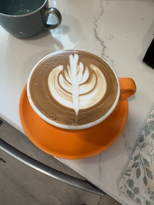 An overhead shot of an orange cup of coffee on a matching saucer, featuring beautiful latte art in a rosetta/leaf pattern, sitting on a white marble table next to a gray mug.