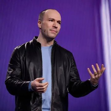 A man wearing a black leather jacket over a light blue shirt stands on a stage against a purple background, gesturing with his hands while speaking.