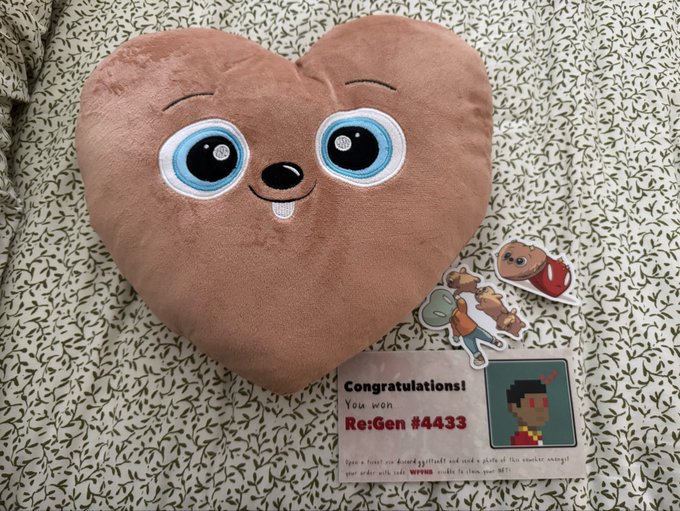 A heart-shaped brown plush pillow with a smiling face rests on a leafy-patterned blanket, accompanied by character stickers and a card reading 'Congratulations! You won Re:Gen #4433' with a Minecraft-style pixel avatar.
