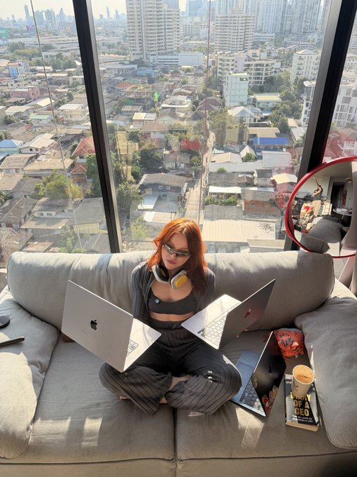 A woman with red hair and glasses sits cross-legged on a gray couch in front of a window overlooking a city, working on two laptops (an Apple MacBook and another laptop), with headphones around her neck and a book titled 'OF A NDEO' beside her.