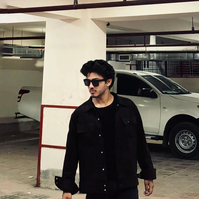 Stylized portrait of a young man with dark hair wearing black sunglasses and a black corduroy shirt, standing in a parking garage with a white pickup truck behind him.