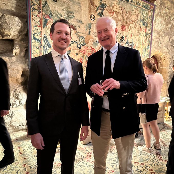 Two men in formal attire pose for a photo in front of an ornate medieval tapestry at an indoor event; the younger man wears a dark suit with a light blue patterned tie, while the older gentleman wears a dark blazer with a black tie and holds a glass.
