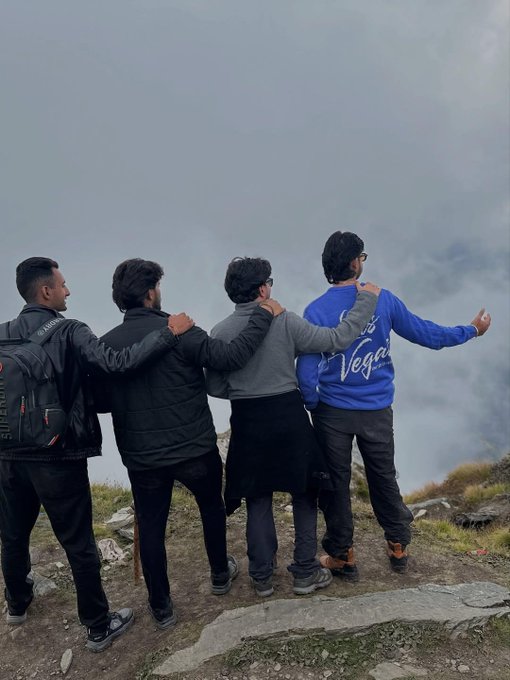 Four young men stand with their arms around each other on a mountain peak, looking out over a sea of clouds.