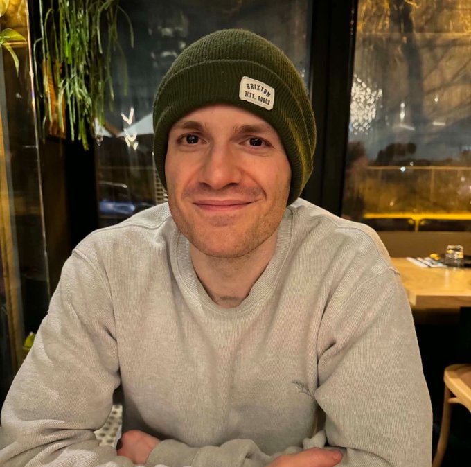 A man wearing an olive green Brixton beanie and a light gray sweatshirt smiles warmly while sitting in what appears to be a restaurant.