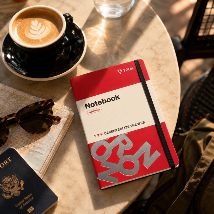 A red and white notebook branded with 'TRON' and 'DECENTRALIZE THE WEB' sits on a table alongside a cappuccino, sunglasses, a US passport, and a map in a lifestyle product photo.