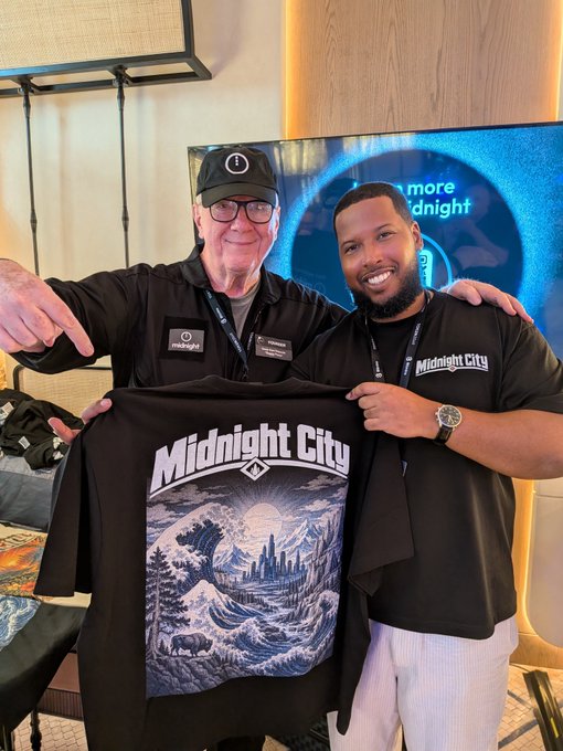 Two men wearing 'Midnight City' branded apparel; they smile while displaying a black t-shirt featuring a 'Midnight City' wave-and-bison graphic design.