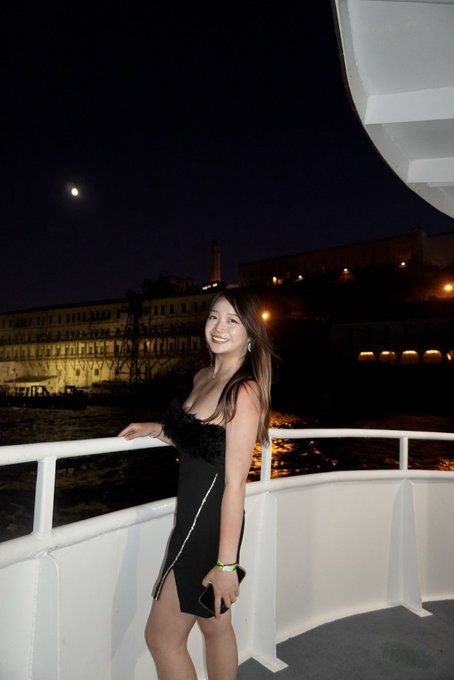 A young woman in a short black dress stands on the deck of a boat at night, smiling at the camera with illuminated waterfront buildings in the background.