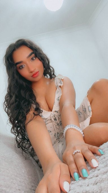 A young woman with long curly dark hair poses on a light carpet wearing a sheer floral dress, featuring turquoise nail polish, a pearl bracelet, and a ring.
