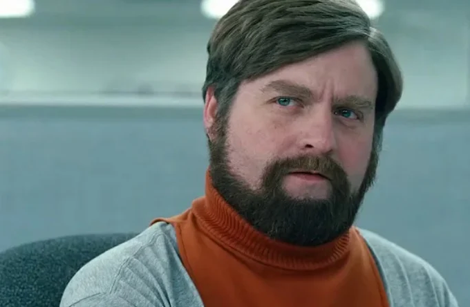 Close-up photograph of actor Zach Galifianakis with a beard, wearing an orange turtleneck under a gray sweater, displaying a serious expression.