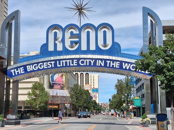 A photograph of the iconic Reno Arch sign reading 'RENO' and 'THE BIGGEST LITTLE CITY IN THE WORLD' spanning over a city street on a sunny day.