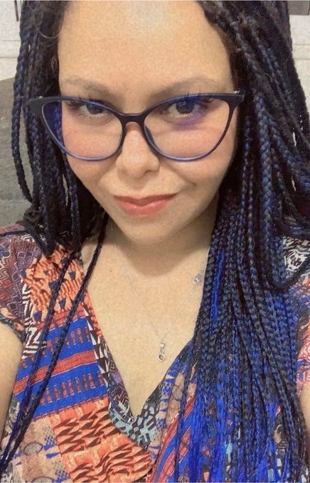A selfie of a woman with long blue braided hair wearing black cat-eye glasses and a colorful patterned top with a silver necklace.