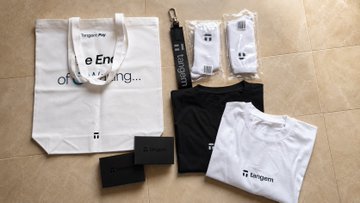 The image features a variety of merchandise items, including a T-shirt, socks, a keychain, a tote bag, and some other accessories. The items are arranged in an unorganized manner, with each item placed next to one another on a tiled floor. The post that posted this image is titled "@Tangem withe socks logo @aigarxyz who sent me this as an ambassador merch This is what I got # Tangem withe socks # Tangem keychain # Tangem Pay white tote bag # Semiconductor smart cards set with Tangem Ring". The post seems to be promoting a product or service, possibly related to the T-shirt and other items in the image.