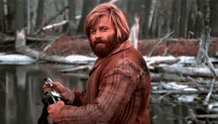 The image features a man with a beard wearing a brown jacket standing near water. He has his arm outstretched towards the camera, holding an object that appears to be a gun or a rifle. The background of the image shows trees and bushes, creating a natural setting for this scene. The post is titled "NOW I AM ALL IN ON XRP."