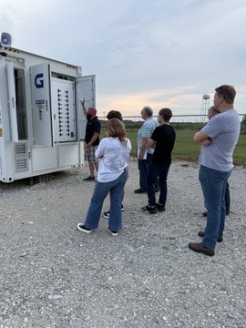 A group of five people gather outdoors around an open electrical cabinet or industrial unit, with one person appearing to point or interact with its contents.