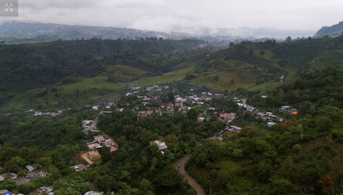 An aerial drone view captures a small village nestled in a green, forested mountain valley under a cloudy sky.