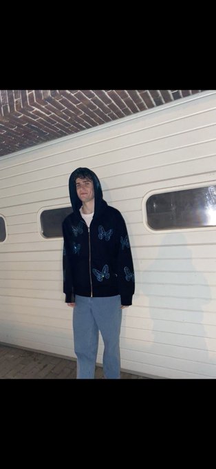 A young man in a dark butterfly-patterned zip-up hoodie and light blue jeans stands against a white paneled wall under a brick archway.