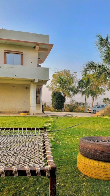 A two-story house with a grassy yard features a rope-woven charpai cot and stacked tire planters in the foreground. The image sets a relaxed, outdoor scene that complements the post's invitation to play the @sleepagotchi sleep game on Telegram, evoking a peaceful environment perfect for rest or gaming.