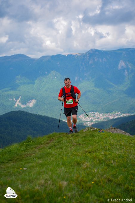The image shows a man hiking up a mountain with his skis attached to his backpack while wearing a red shirt and black shorts. The man appears to be enjoying the outdoors, as he takes breaks during his hike to appreciate the beautiful surroundings. The photo is taken from an elevated perspective, providing a clear view of the mountains in the background.