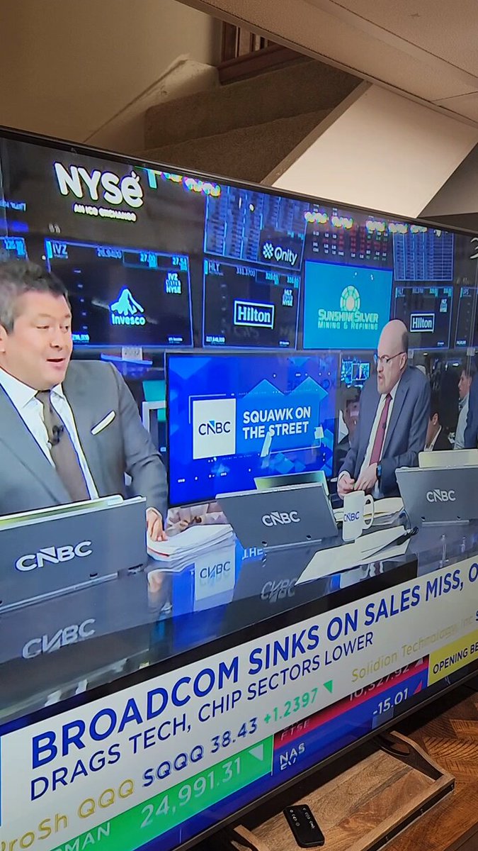 In this image, a man sits at a desk with an electronic television screen displaying the stock market news. On the TV, two men can be seen sitting behind computer monitors, likely working on analyzing the data or sharing their insights about the market. The background of the scene features many screens and logos for various companies such as Crain's, Dow Jones & Co., and Broadcom. Additionally, there is a reference to the Bitcoin exchange rate multiple times in different descriptions within this post. This suggests that the focus of the image is on the stock market discussion happening on television, highlighting the ongoing interest and engagement around financial news and its impact on the market.