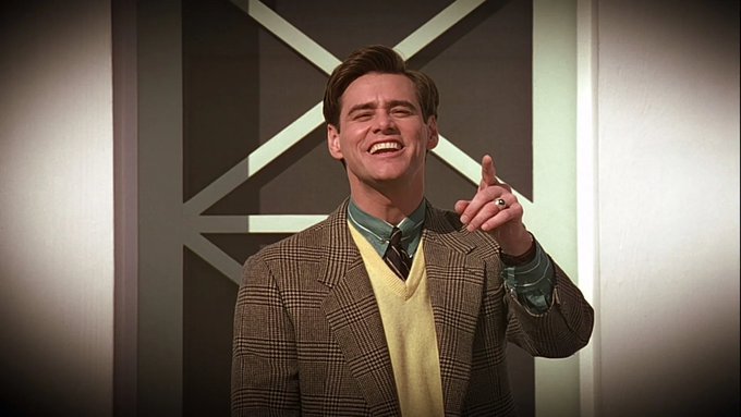 A smiling man in a brown plaid blazer with a yellow sweater vest points upward in a vintage-style room.