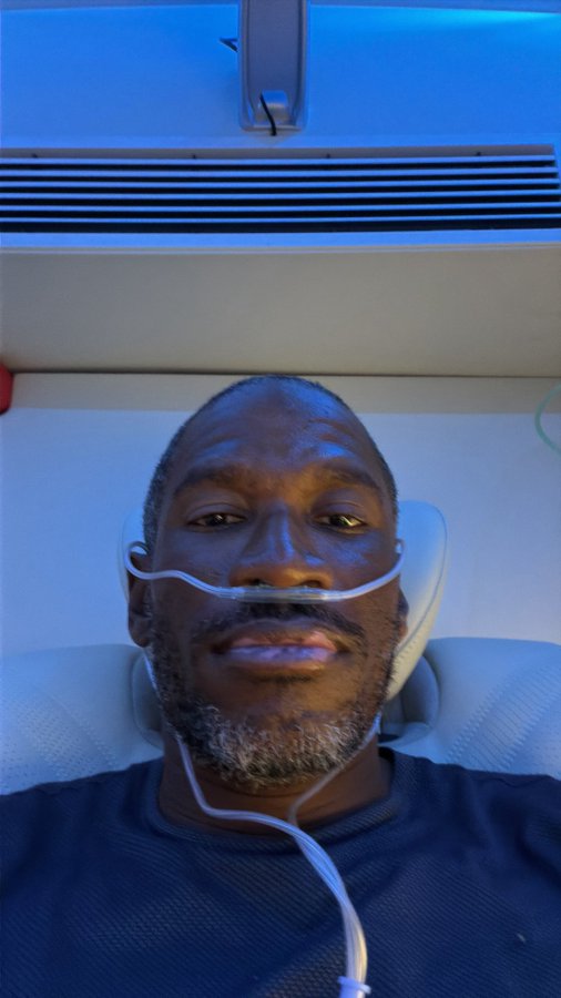 A selfie of a man receiving medical treatment, wearing a nasal cannula while lying in a chair under blue-lit equipment.