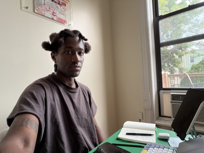 A young man with short dreadlocks sits at a desk by a window in a room, looking toward the camera with a computer and notebook in front of him.