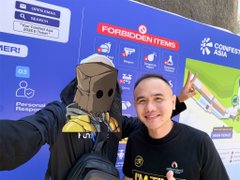 A man in a black shirt poses with a person wearing a traditional hooded costume at a Crypto Asia conference, with a "Forbidden Items" sign visible behind them. The image relates to the post as a visual introduction to Mr. Gema, whose expertise in trading and market analysis is discussed in the interview.