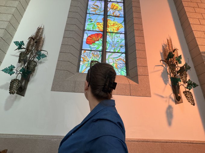 A woman with dark hair in a bun views a stained glass window depicting flowers and dragonflies inside what appears to be a church or chapel, flanked by decorative metal wall sconces.