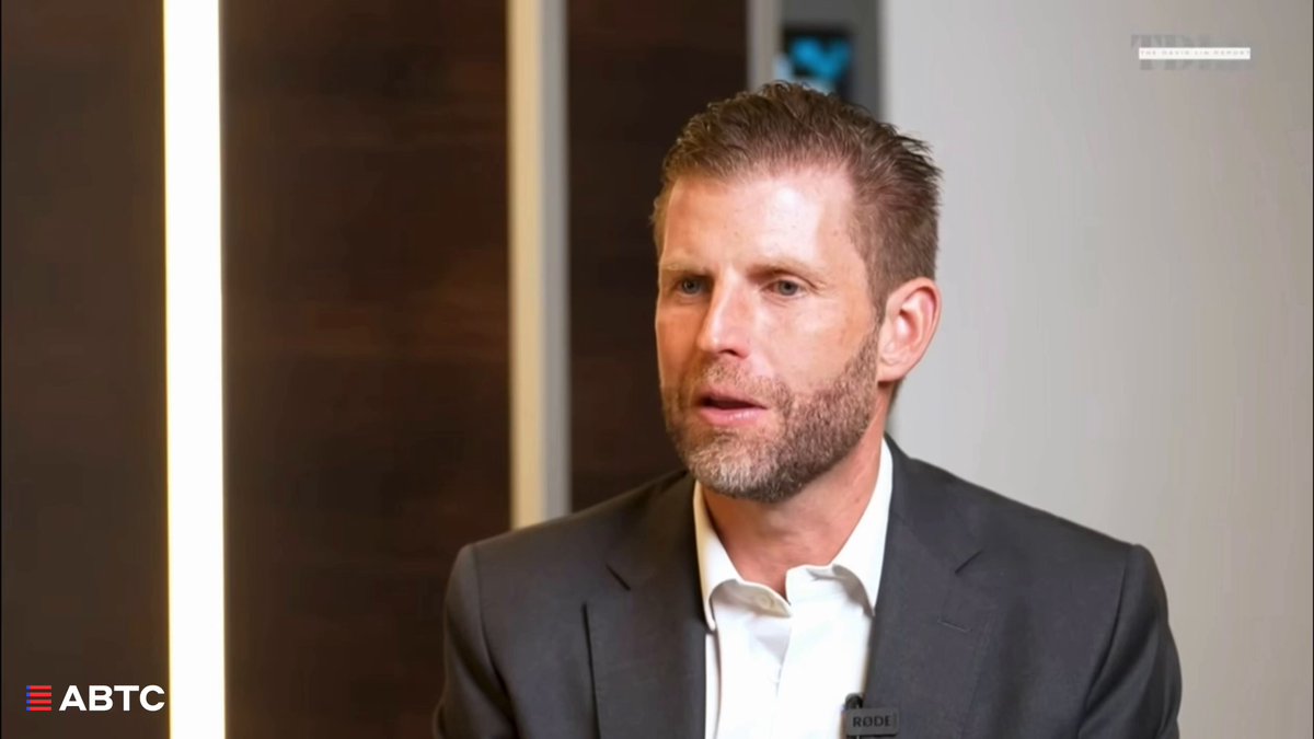The image shows a man wearing a suit, standing against a white wall with a wooden door behind him. The man appears to be giving a speech or presentation, as indicated by his body language and facial expressions. The focus of the image is on the man in the suit, capturing his attention and engagement during the event.