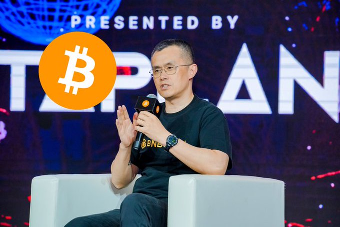 The image features a man sitting in a chair, holding a microphone and speaking into it. He appears to be giving a speech or presentation related to Bitcoin. The background of the image shows a large screen displaying the words "Bitcoin" written on it. The man is wearing glasses and has a serious expression as he speaks about Bitcoin's future.
