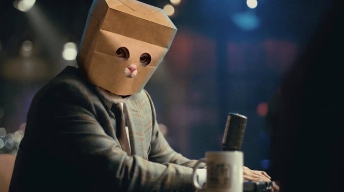 A person wears a cardboard box over their head with eye and mouth holes cut out while sitting at a microphone in a dimly lit setting.