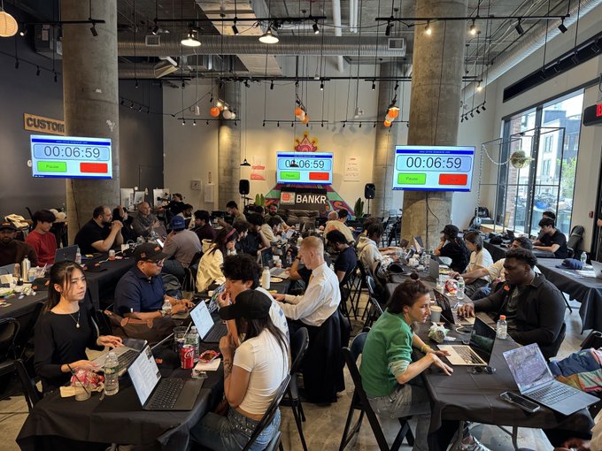 A large room filled with people working on laptops at tables, with countdown timers displayed on screens reading '00:06:59' and a 'BANKR' logo visible.