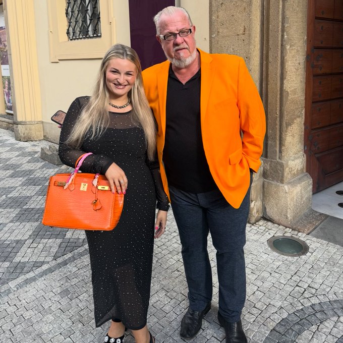 A man in a bright orange blazer stands beside a woman in a black dress holding a matching orange Birkin bag, posing together outside a building. The man's vibrant attire and the setting suggest this is a memorable occasion marking the transition from one chapter to the next in his life.
