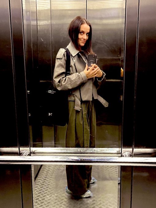 A woman stands in an elevator taking a mirror selfie with her phone, wearing a gray jacket, green wide-leg pants, and carrying a black bag, with the caption "I'm 40 btw" playfully highlighting her youthful appearance. The metallic elevator doors and floor create a sleek backdrop for the casual, confident pose.