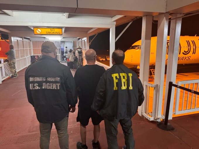 An FBI agent and a Diplomatic Security U.S. agent escort a man through an airport gate at night, with a passenger plane visible. The scene reflects the reported federal action in a $165 million crypto-investment scheme that allegedly defrauded more than 6,000 investors through promised monthly returns of 25%.