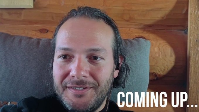 A man with long dark hair and a beard speaks into a wired earphone, with 'COMING UP...' text overlaid on a wooden interior background.