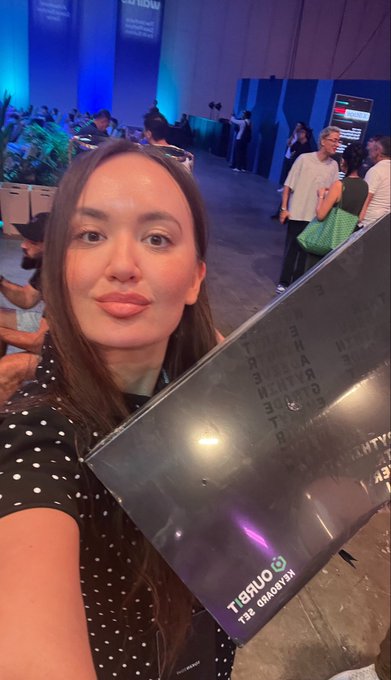 A woman in a black and white polka dot top takes a selfie while holding a large award folder with 'TikTok' branding, inside a convention hall or awards venue.