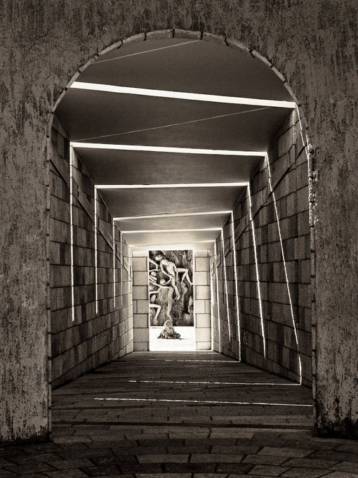 A black and white photo of a brick archway looking down a corridor lined with illuminated geometric shapes towards an elephant statue.
