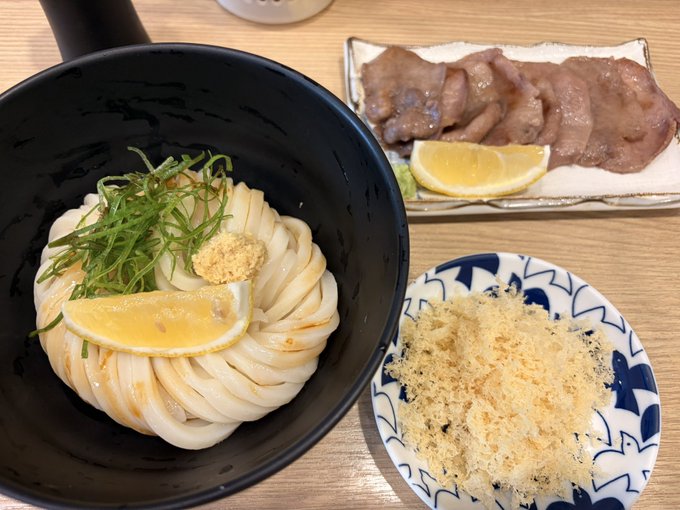 A Japanese meal consisting of a bowl of udon noodles topped with a lemon slice, ginger, and green onions, accompanied by a plate of sliced beef with lemon and a small dish of katsuobushi (fish flakes).
