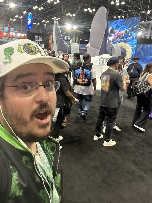 Selfie of a man with glasses and a white cap at what appears to be an anime convention, with large character props and attendees visible in the background.