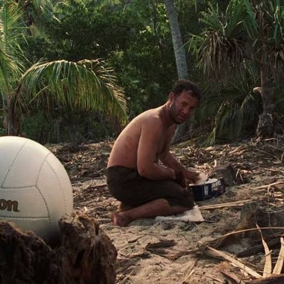 A shirtless, bearded man sits on a sandy beach near palm trees, cooking over a small fire beside a large white volleyball with partial text reading "on" visible. The caption referencing CZ (Binance CEO Changpeng Zhao) humorously compares the rugged survival scene to crypto traders weathering market "shaking," suggesting that like the stranded castaway, holders will outlast attempts to be "shaken out."