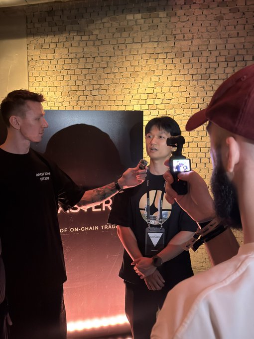 A man in a black shirt is being interviewed at what appears to be a crypto event, with text visible on a backdrop mentioning 'ON-CHAIN TRADING' and 'INVESTICON EST 2018'. Another person with a red cap is filming the interview with a camera.