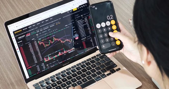 An over-the-shoulder view of a person using a calculator app on a smartphone while looking at Binance trading charts on a MacBook laptop.