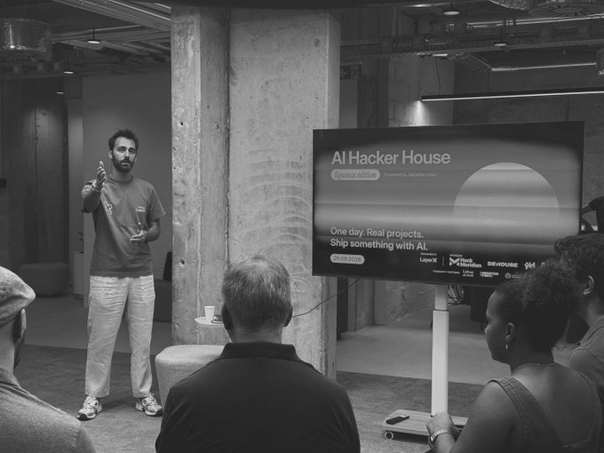 A black-and-white photo captures a man presenting at the 'AI Hacker House' event (Dayton edition), with a screen displaying 'One day. Real projects. Ship something with AI.' and sponsor logos including Logyx, Hack Mobb, and D5 HOUSE.